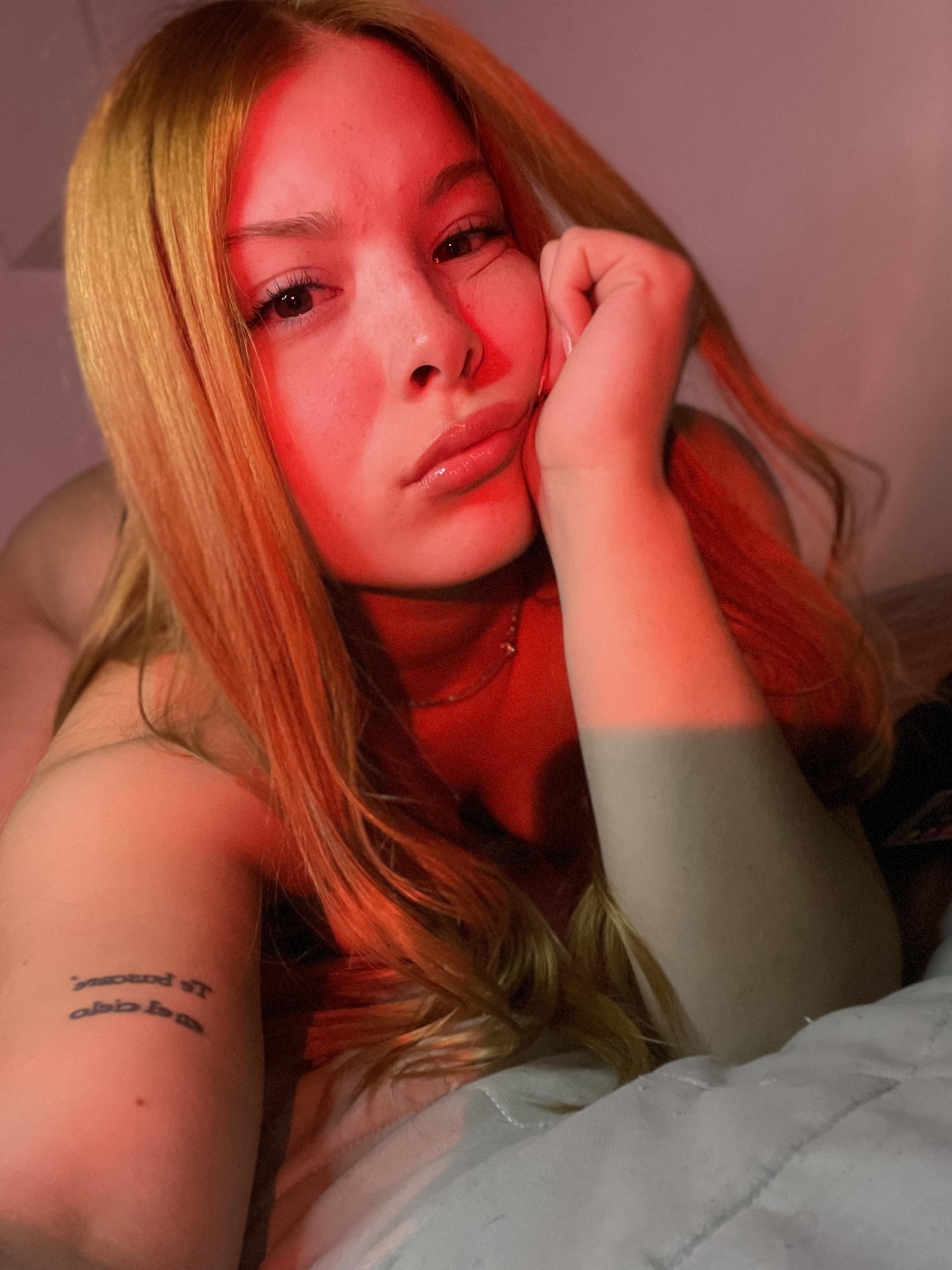 Mia Prescott's red-lit face, hand on cheek, long blonde hair, and tattooed arm, anticipating your touch.