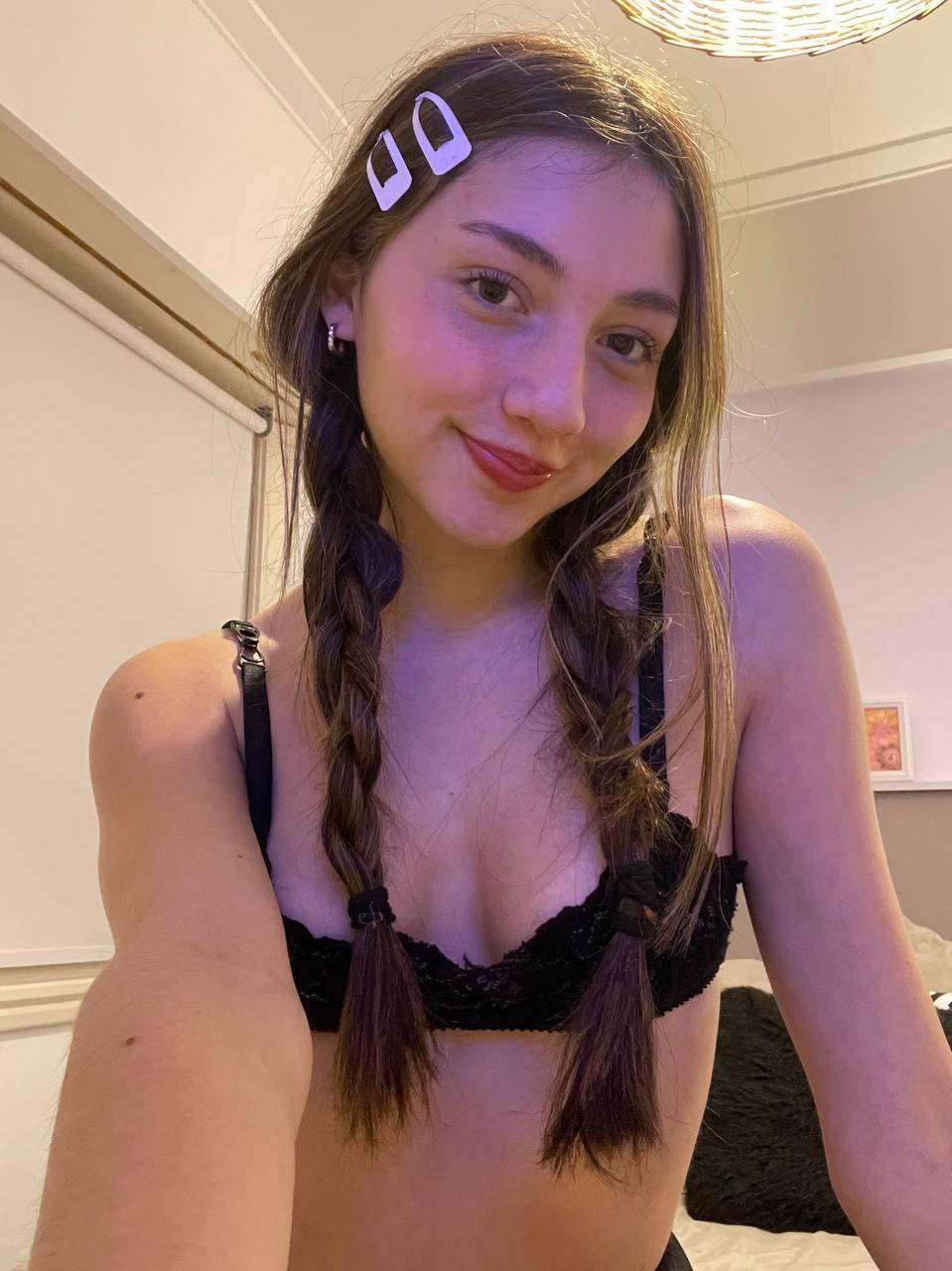 Kiera Brooks, with braided hair, leans forward, her black bra teasing her small, perky breasts, smiling seductively.