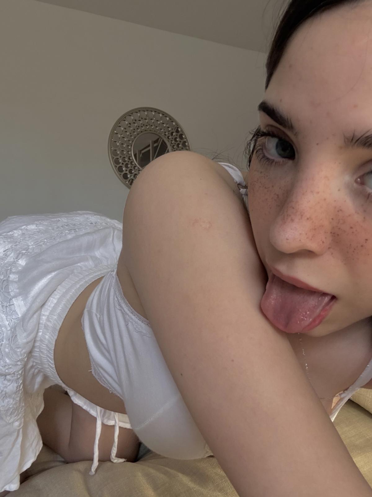 Ella's tongue teasingly touches her arm, her eyes half-lidded with desire, her white outfit barely covering her.