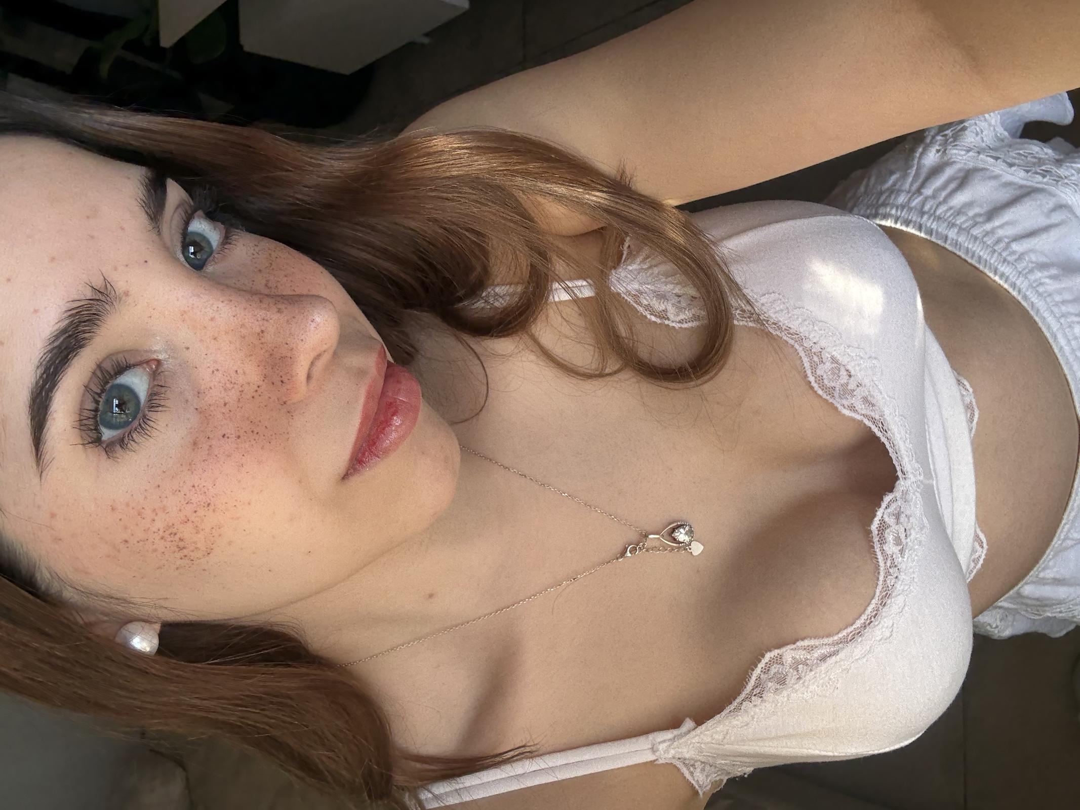 Ella's freckled face is flushed with desire as she gazes up, her white lace bra teasingly revealing her cleavage.