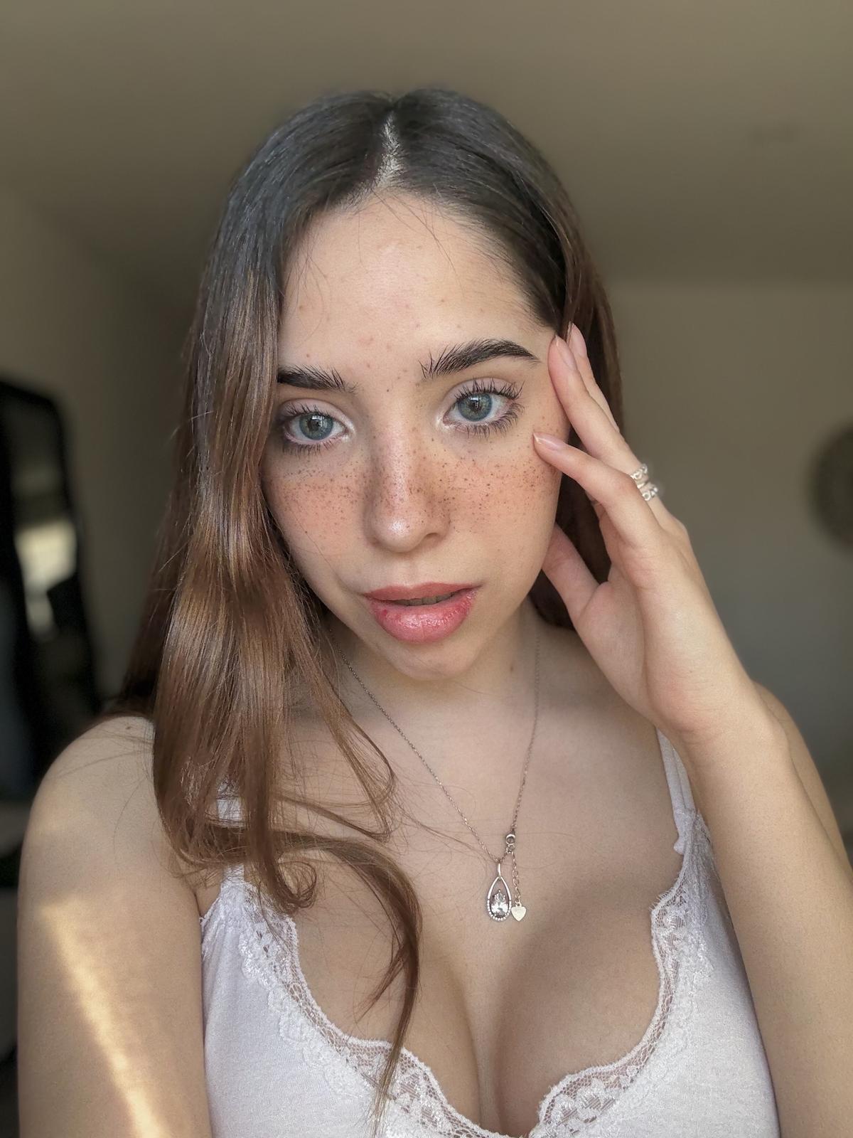 Ella's hand touches her face, her freckled skin glowing, her cleavage inviting, heart necklace resting against her chest.