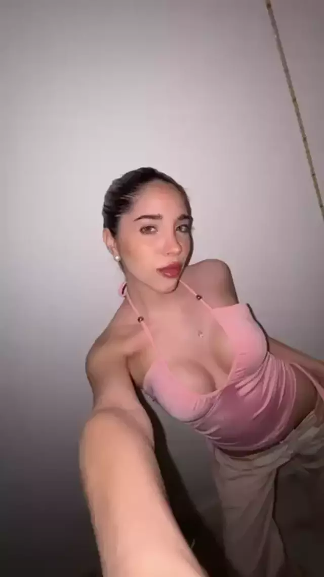 Ella's pink halter top accentuates her cleavage, her hand reaching out, teasing with a seductive, inviting smile.